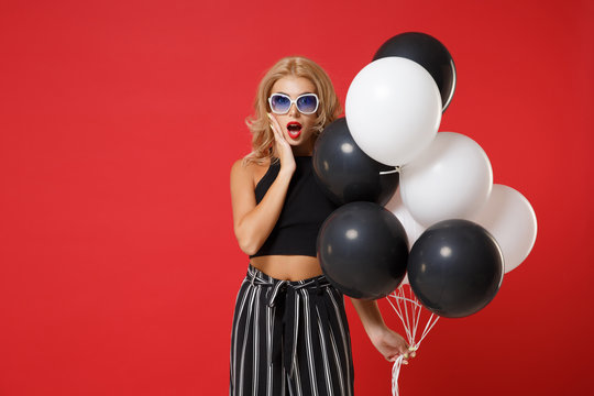 Shocked woman in black clothes, glasses posing isolated on red background. Valentine's Day, birthday holiday party concept. Mock up copy space. Celebrating holding air balloons, keeping mouth open.