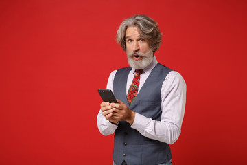 Shocked elderly gray-haired mustache bearded man in classic shirt vest and colorful tie posing isolated on red background in studio. People lifestyle concept. Mock up copy space. Holding mobile phone.