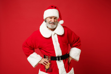 Fototapeta na wymiar Elderly gray-haired mustache bearded Santa man in Christmas hat posing isolated on red background. Happy New Year 2020 celebration holiday concept. Mock up copy space. Stand with arms akimbo blinking.