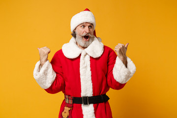 Fototapeta na wymiar Shocked elderly gray-haired bearded mustache Santa man in Christmas hat posing isolated on yellow wall background. New Year 2020 celebration holiday concept. Mock up copy space. Pointing thumbs aside.
