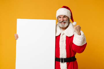 Fototapeta na wymiar Elderly gray-haired mustache bearded Santa man in Christmas hat isolated on yellow background. New Year 2020 celebration concept. Mock up copy space. Hold white blank billboard, doing winner gesture.