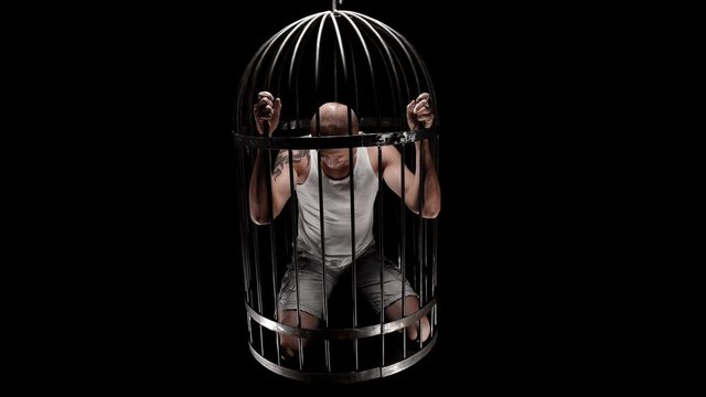 a person in a bird&rsquo;s cage is held captive, raises his head and looks into the camera, maybe each of us is locked in his own cage 4K