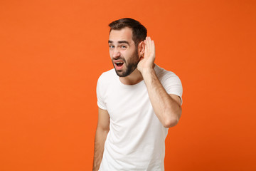 Fototapeta na wymiar Concerned young man in casual white t-shirt posing isolated on orange background, studio portrait. People sincere emotions lifestyle concept. Mock up copy space. Try to hear you with hand near ear.