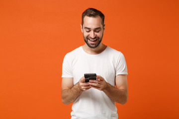 Smiling young man in casual white t-shirt posing isolated on orange background, studio portrait. People sincere emotions lifestyle concept. Mock up copy space. Using mobile phone, typing sms message. - obrazy, fototapety, plakaty