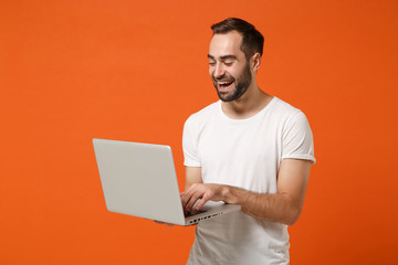 Laughing young man in casual white t-shirt posing isolated on bright orange background studio portrait. People sincere emotions lifestyle concept. Mock up copy space. Working on laptop pc computer.
