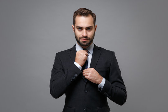 Confident young business man in classic black suit shirt tie posing isolated on grey wall background studio portrait. Achievement career wealth business concept. Mock up copy space. Straightening tie.