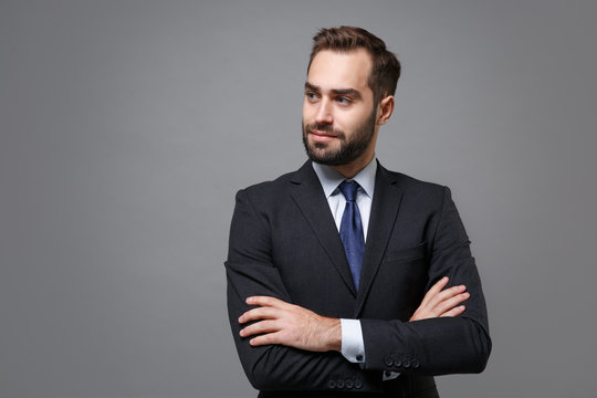 Handsome young business man in classic black suit shirt tie posing isolated on grey background. Achievement career wealth business concept. Mock up copy space. Holding hands crossed, looking aside.