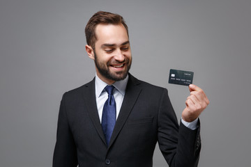 Smiling young business man in classic black suit shirt tie posing isolated on grey wall background in studio. Achievement career wealth business concept. Mock up copy space. Holding credit bank card.