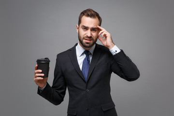 Tired young business man in classic black suit shirt tie posing isolated on grey background. Achievement career wealth business concept. Mock up copy space. Hold cup of coffee or tea put hand on head.