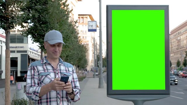 On a city street next to a large billboard green screen stands a caucasian middle aged man and clicks into a smartphone smiling in the background cars are driving people are passing a Summer Sunny day