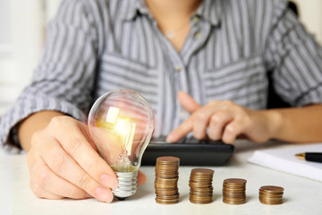 Woman with light bulb, calculator and coins at white table, closeup. Power saving