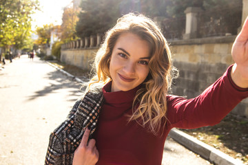 Close up sunny portrait of young happy pretty girl making selfie and smile to the camera, wearing a burgundy dress and a checkered autumn coat, with blond curly hair.