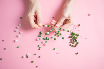 Women's hands make crafts from white and green beads. Pastel pink background. The concept of creativity, handwork, preparation for the holiday. Minimalism, top view, flat lay.