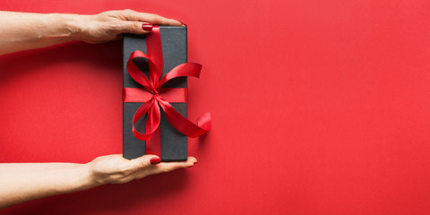 Woman's hands holding black gift box wrapped with red ribbon on red surface.