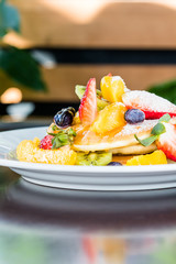 Pancakes on a white plate, decorated with fruits: strawberries, mangoes, orange, kiwi and blueberries and drizzled with natural honey and sprinkled with powdered sugar.Pancakes on a white plate, decor