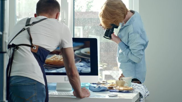 Handheld medium shot of man with ginger hair holding digital camera and taking pictures of food lying on tablecloth as male cook in apron looking at photos on computer screen