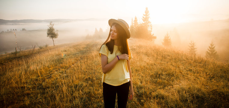 Carefree happy woman in yellow shirt and straw hat enjoying nature on grass meadow on top of mountain with sunrise. Beauty girl outdoor with sunbeams. Freedom concept