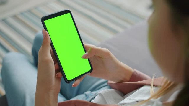 Close-up of woman using smartphone with blank chroma key screen in house sitting alone swiping enjoying social media online. People and internet concept.