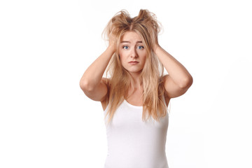 Young woman in white shirt disappointed with her messy and disheveled blond hair, standing with her arms to her head, looking at camera with sad emotions. Front portrait against white background