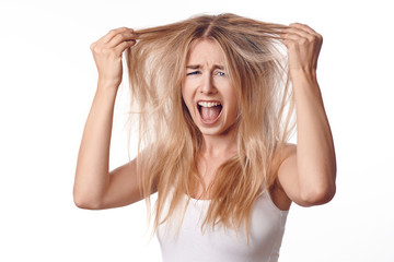 Young blond woman unhappy with her haircut, touching her hair with both hands and looking at camera with screaming grimace. Front portrait on white background - obrazy, fototapety, plakaty
