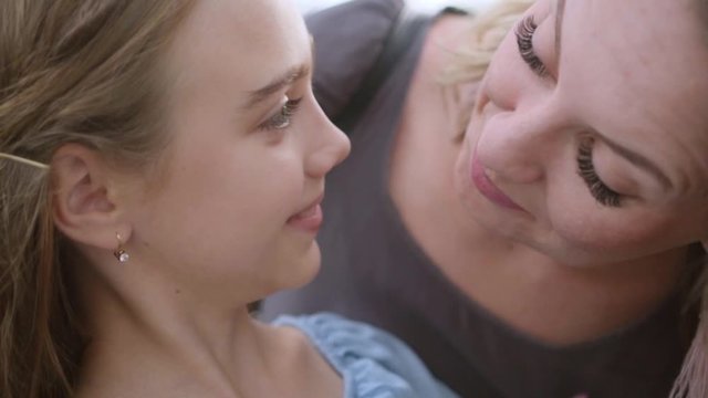 mother with false eyelashes, in brown dress leans to her daughter, the long-hair 10 years girl in blue smiles broadly in response, close-up. wrinkles on the mom s forehead