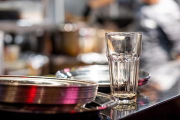 A tall empty glass water glass and a stack of plates on a table in a restaurant. Heavily blurred background