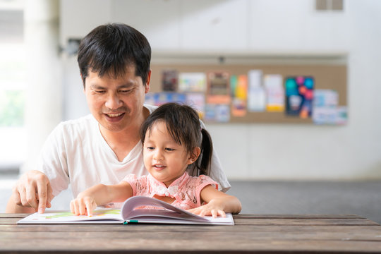 Asian Father And Daughter Are Reading The Storybook Together And They Are Smiling With Happy Moment, Concept Of Learning Activity For Kid In Family Lifestyle. And Role Of Parent Support To The Child.