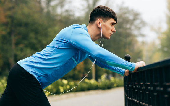 Side view image of young sportsman stretching outdoor in the morning on the fog nature background. Fitness male exercising in the park and listenting the music on earphones. People and sport concept