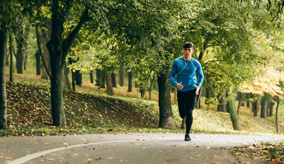 Horizontal full body image of young jogger man jogging outdoor in the forest background. Fitness male exercising in the park, wearing blue and black sportswear. People and sport concept © Z A C