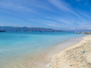 Lots of boats anchored next to the shore of Gili Air, Lombok Indonesia. Beautiful and clear water. In the back visible Mount Rinjani. Some trees on the shore, few clouds on the sky. Holidays paradise