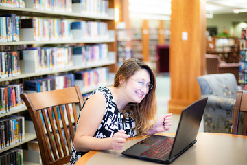 White female, Caucasian woman sitting at the desk with laptop.  she is happy, smiling and looking on computer. Library or bookstore