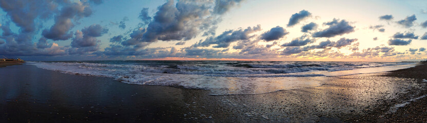 Awesome panoramic 180 degrees of rough sea at sunset on the beach, a dramatic sky with amazing light and cloudscape and sun reflections with on water edge crashing