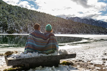 Back view of a man and woman couple covered with a tribal blue blanket sitting on a stone bench while are looking at a beautiful icy lake surrounded of snowy forest and mountains. Winter wonderland.