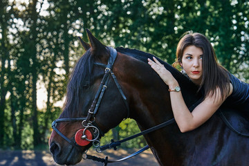 Young slim woman brunette in a black dress sitting on dark brown horse. Sunny summer evening. Close-up.