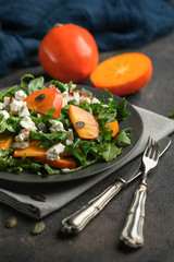 Healthy salad with persimmons, arugula, feta cheese and pumpkin seeds on a dark plate on a dark background. Vegetarian menu, healthy food. Prevention of vitamin deficiency.