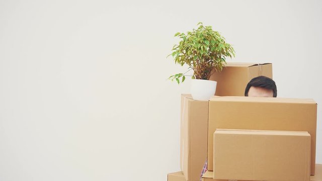 Funny young man is appearing from behind the stack of cardboard boxes, looking around, raising eyebrows, showing keys, disappearing.