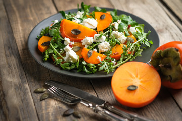 Healthy salad with persimmons, arugula, feta cheese and pumpkin seeds on a dark plate on a dark background. Vegetarian menu, healthy food. Prevention of vitamin deficiency.