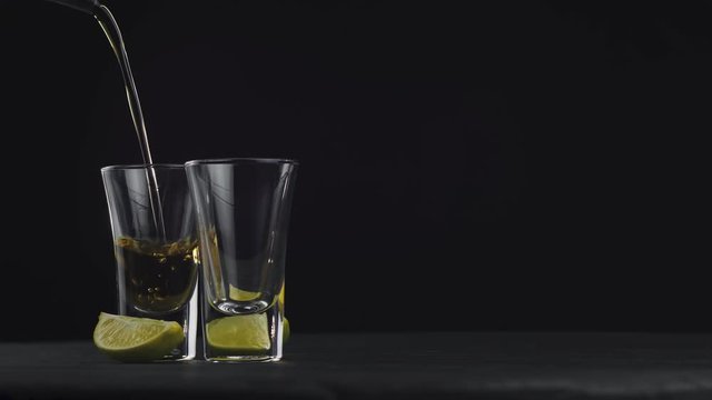 Pouring golden tequila in two glasses on a black background on a rotating black board. Pour of alcohol drink vodka tequila. Close-up shot. . 4k video. 59.94 fps