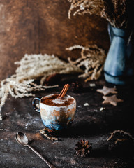 mug of coffee with cinnamon stick on brown table with cookies, dry herbs and pine cones, festive card