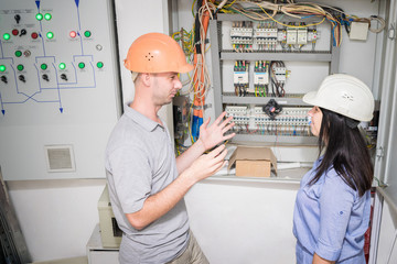 Obraz premium A team of electricians are talking near the electrical panel. A man and a woman in engineering helmets discuss the power supply of the enterprise. Specialists work in the electric box.
