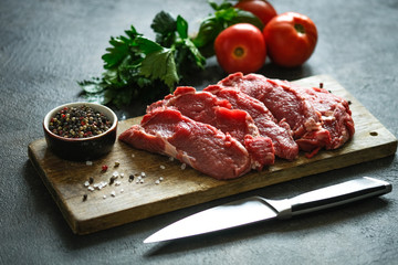 Fresh raw beef, cut into steaks on a wooden board with vegetables, herbs and spices on a wooden board on a dark background.