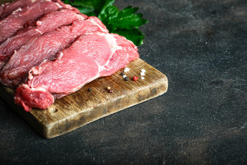 Fresh raw beef, cut into steaks on a wooden board with vegetables, herbs and spices on a wooden board on a dark background.