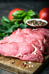 Fresh raw beef, cut into steaks on a wooden board with vegetables, herbs and spices on a wooden board on a dark background.