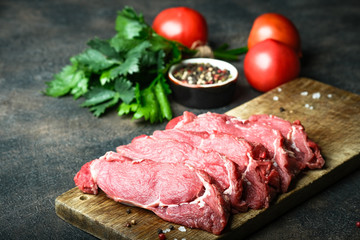 Fresh raw beef, cut into steaks on a wooden board with vegetables, herbs and spices on a wooden board on a dark background.