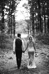 Fototapeta premium Black and white foto the newlyweds walk hand in hand along the path in the forest. Back view. Bride and groom enjoying romantic moment together on wedding day. The concept of youth, love and lifestyle