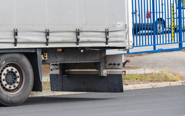 rear of the trailer of a gray truck parked at the door of a factory with a view of the fence and a van inside, the road and a safety pole