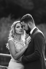Naklejka premium Black and white foto stylish couple of newlyweds posing on a bridge on wedding day. Handsome bearded groom admires and kisses pretty bride. Together. The concept of youth, love, fashion and lifestyle.