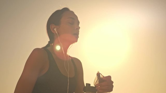 Tired young fit sporty woman with lip piercing wearing a black tracksuit and wired headphones drinks some water from sport bottle then watering herself during training outside in the morning
