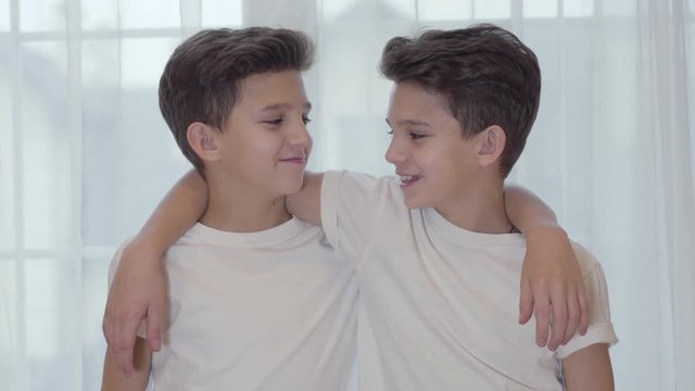 Portrait of Caucasian twins looking at camera and smiling. Twin brothers in similar white T-shirts keeping hands on each other's shoulder and talking. Siblings posing indoors.