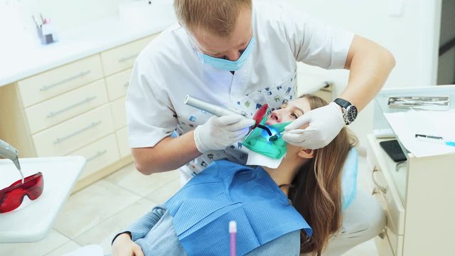 A male dentist in white latex gloves polymerizes a filling using a device with UV light. A girl treats teeth for caries in the dentist's office.
